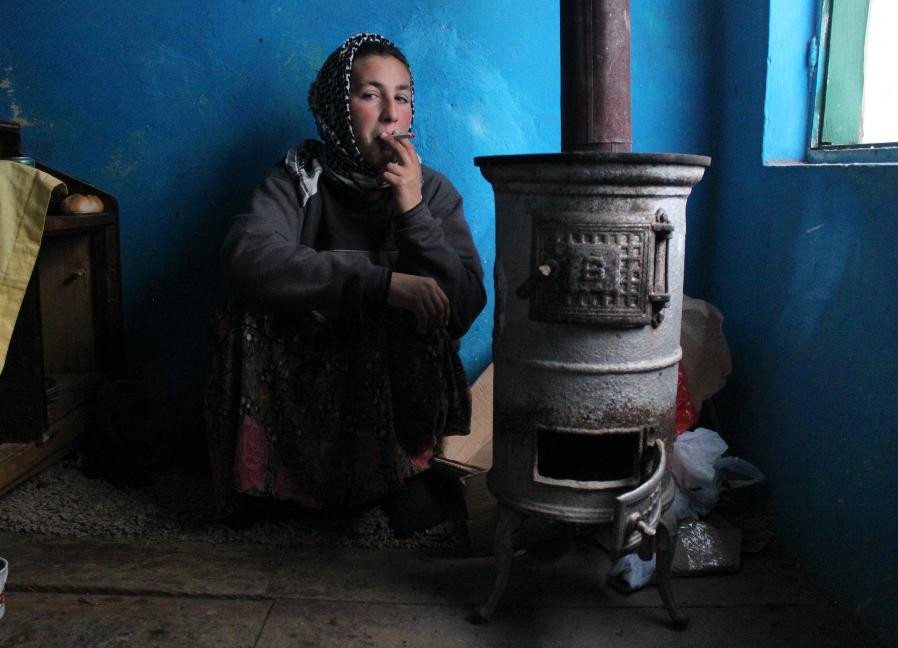 A young Roma women crouches beside the wood stove in her house while smoking a cigarette. She wears a scarf over her head, a long skirt, and a bulky sweater.