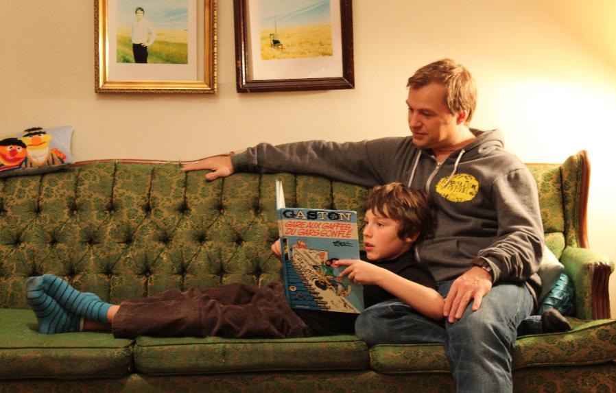 A young boy lays on the couch reading a French-language picture book while his father sits behind him smiling.