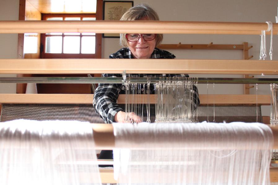 A woman smiles through wooden slats of her loom. She weaves white strands of yarn.